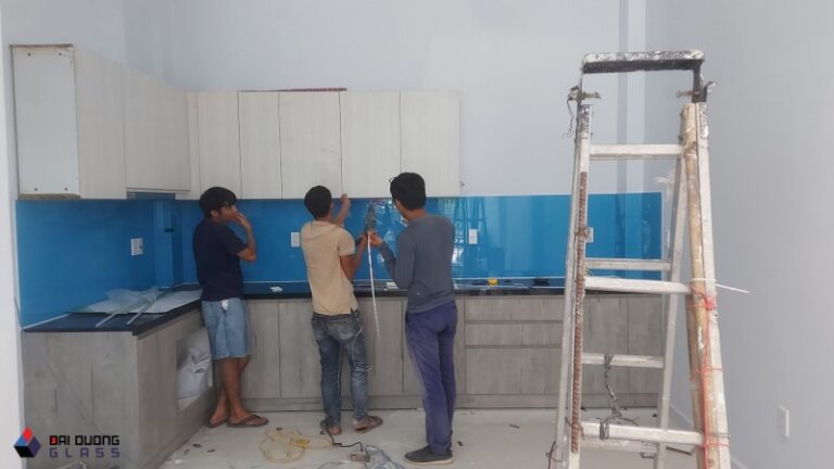Ba thợ lắp kính ốp bếp màu xanh dương lên tường bếp, cạnh thang nhôm